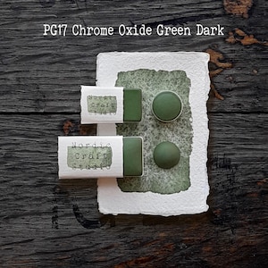 Könnte beinhalten: Zwei rechteckige Näpfchen mit Chrome Oxide Green Dark Aquarellfarbe, beschriftet mit PG17, jeweils in einer weißen Papierhülle mit dem Text Nordic Craft Studio, neben zwei runden grünen Farbmustern auf einem strukturierten weißen Papier.