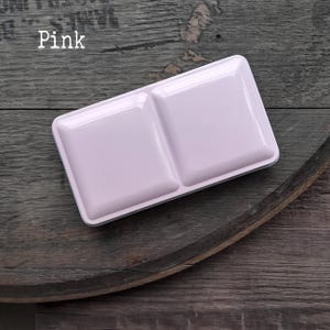 Könnte beinhalten: Ein rechteckiger, hellrosa Aquarellfarbkasten aus Metall mit zwei großen, erhabenen, quadratischen Fächern auf dem Deckel. Das Wort Pink ist in weißer Schrift in der oberen linken Ecke des Bildes über dem Farbkasten aus Metall aufgedruckt.