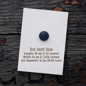 Puede incluir: Una tarjeta rectangular blanca con un punto circular azul oscuro. El texto dice: Dot Card Size, Length: 89 mm, Width: 64 mm, Dot Diameter: 14 mm. La tarjeta tiene una superficie texturizada y se encuentra sobre una base de madera oscura.