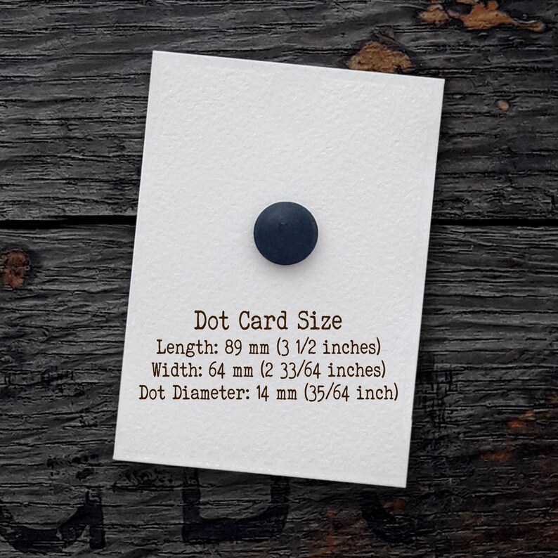 Puede incluir: Una tarjeta rectangular blanca con un punto circular azul oscuro. El texto dice: Dot Card Size, Length: 89 mm, Width: 64 mm, Dot Diameter: 14 mm. La tarjeta tiene una superficie texturizada y se encuentra sobre una base de madera oscura.
