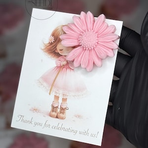 Puede incluir: Tarjeta de felicitación con una ilustración en acuarela de una niña con un vestido rosa y una gran flor rosa adjunta. La tarjeta dice "Thank you for celebrating with us!" en una fuente de escritura.