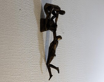 May include: A bronze-colored wall sculpture depicting two figures in a geometric, abstract style. One figure appears to be helping the other climb. The sculpture is mounted on a textured, off-white wall.