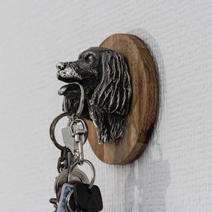 May include: A key holder featuring a dog head sculpture made of metal, mounted on a round wooden plaque. The dog's head is detailed with fur and facial features. Several keys hang from the key holder.