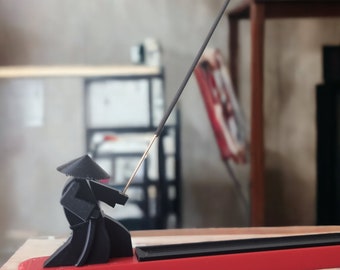 Puede incluir: Un porta incienso negro estilo origami con forma de figura de samurái, sosteniendo un palito de incienso. El soporte se asienta sobre una base rectangular roja. El palito de incienso es gris y tiene una punta dorada. El fondo está borroso.
