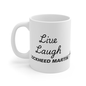 Op de afbeelding: Witte keramische mok met een gebogen handvat. De mok heeft de woorden "Live Laugh" in een cursief lettertype, met "Lockheed Martin" eronder gedrukt in een schreefloos lettertype.