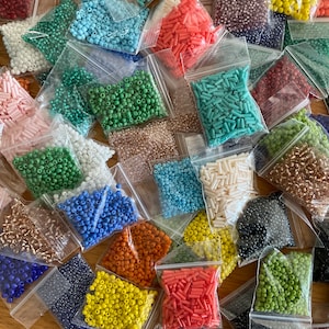 Może przedstawiać: Zbiór małych plastikowych torebek wypełnionych koralikami w różnych kolorach. Koraliki mają różne kształty i rozmiary, w tym okrągłe, kwadratowe i cylindryczne. Torebki są ułożone w przypadkowy wzór na drewnianej powierzchni.