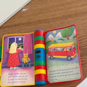 May include: A colourful children's musical book featuring two open pages. The left page shows a person looking out a window at night with a teddy bear and the text Twinkle, twinkle, little star. The right page shows a red bus and the text The wheels on the bus go round and round. The spine has blue, green, yellow, and red buttons.