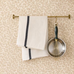 May include: A brass-toned metal rail holds a white kitchen towel featuring a single vertical dark charcoal stripe. A stainless steel whisk with a dark green handle hangs from the rail by a matching brass-toned metal hook next to the towel.