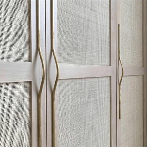 Peut inclure: Une armoire en bois clair dotée de plusieurs portes avec des panneaux en cannage tressé et de longues poignées en laiton incurvées et verticales. Le cadre de l'armoire a une finition lisse et pâle, et les inserts des portes présentent une texture en maille fine.