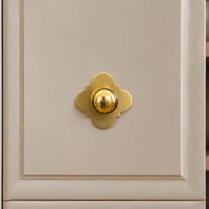 May include: A gold-toned metal cabinet knob featuring a central spherical protrusion mounted on a flat, four-lobed clover-shaped backplate. The hardware is attached to a light beige cabinet door with a recessed panel frame design.