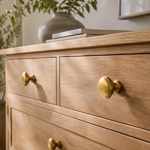May include: A light oak wood chest of drawers featuring a horizontal rectangular frame with two top drawers and a lower drawer. Each drawer is fitted with a round, brushed brass metal knob. The wood grain is visible across the flat drawer fronts.