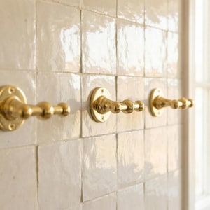 May include: Three brass-colored wall hooks with a decorative design are mounted on a white tiled wall. Each hook has a circular base and a series of rounded, ball-like features extending outward. The hooks are evenly spaced and appear to be made of metal.