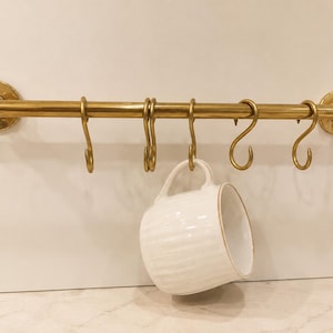 May include: A horizontal brass rail mounted with two circular wall brackets, featuring five matching brass S-hooks suspended from the rod. A single white ceramic mug with a ribbed texture and a gold-toned rim hangs from one of the hooks.