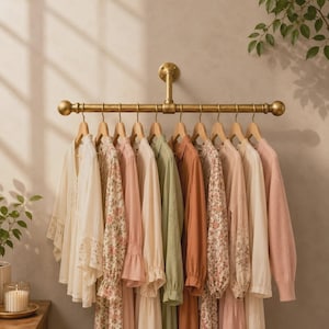 May include: A wall-mounted brass-toned metal clothing rail with a pipe-inspired design, holding ten garments on hangers. The clothing includes a variety of long-sleeved shirts and dresses in neutral, floral, green, orange, and soft pink tones.