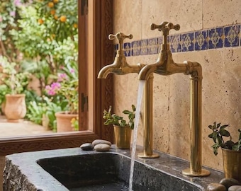 May include: A brass faucet with two cross-handle knobs mounted on a dark, rough-hewn stone sink. Water flows from one spout into the basin. The sink is set against a beige tiled wall with a decorative blue border, featuring small metal pots.