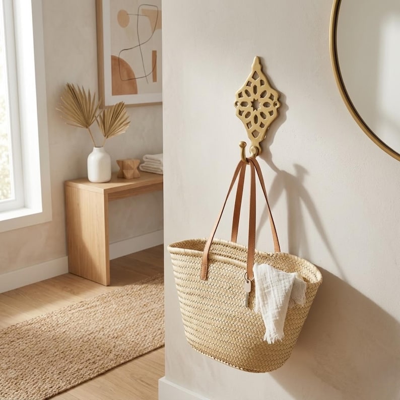 Puede incluir: Un gancho de pared de metal dorado con un intrincado dise&ntilde;o geom&eacute;trico calado montado en una pared. Un bolso de paja trenzada con asas de cuero color canela cuelga del gancho, con una peque&ntilde;a llave plateada y un pa&ntilde;o de tela blanca en una asa.