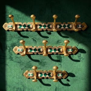 May include: Three brass wall-mounted coat racks of varying lengths, each featuring a series of upward-curving hooks and rectangular inlays with a geometric pattern of dark green and white diamonds set within the metallic frame.