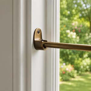 May include: A brass curtain rod bracket mounted to a white window frame with a single dark screw. The bracket holds a horizontal brass rod that extends across the window. The metal has a smooth, matte finish with visible warm golden tones.