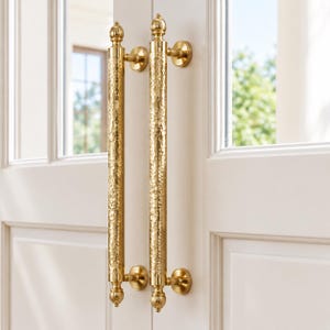 May include: A pair of vertical gold-toned door handles with a textured, hammered finish. Each handle features a cylindrical body with rounded finials at the top and bottom, secured to a white door by two circular mounting plates.