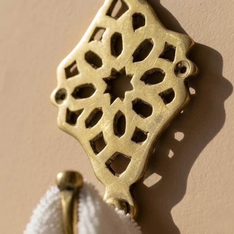 Puede incluir: Un gancho de pared de lat&oacute;n con una placa decorativa que presenta un intrincado patr&oacute;n geom&eacute;trico de estrella calado. El gancho tiene un brazo de metal curvado que sostiene un objeto de tela blanca texturizada, posiblemente una toalla.