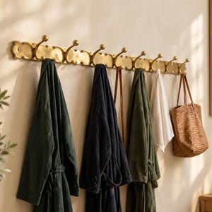 May include: A long, horizontal brass wall-mounted coat rack featuring eight curved hooks with rounded ends. The rack has a scalloped, decorative edge and is mounted to the wall, holding several robes, a white cloth, and a woven straw tote bag.