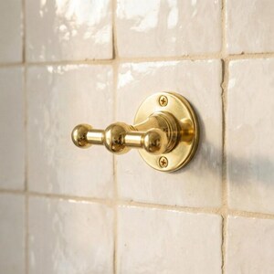 May include: A gold-colored, double-hook wall-mounted fixture. The fixture has a circular base with three visible screws and a horizontal bar with two spherical hooks. The fixture is mounted on white square tiles.