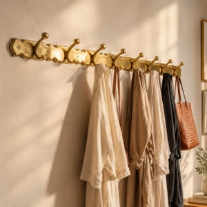 Puede incluir: Un perchero horizontal largo de latón con ocho ganchos curvos, cada uno rematado con una pequeña esfera. El perchero está montado en la pared y sostiene varias prendas colgadas, incluyendo batas de colores claros y un bolso oscuro trenzado.