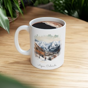 May include: White ceramic mug with a watercolor illustration of a snowy mountain scene with a skier in the foreground. The text "Aspen, Colorado" is printed on the bottom of the mug.