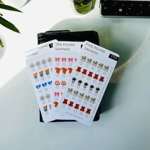 May include: Three saving challenge cards with illustrations of flowers, bows, teddy bears, and cakes. The cards are labeled "Twenty Pound Savings", "Ten Pound Savings", and "Five Pound Savings". The cards are on top of a black wallet. The text "£125" is visible.