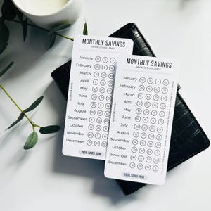 May include: Two white Monthly Savings Challenge cards on a black wallet. The cards feature a savings tracker with circles for each month. Text includes "Monthly Savings" and "Total Saved: £600".