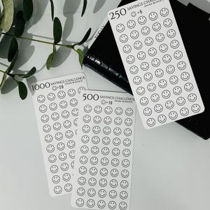 May include: Three white sheets of paper with black smiley face stickers. The sheets are labeled with the text "Savings Challenge" and the amounts "1000", "500", and "250". Each sheet also includes the text "Javan Budgets" and the value of each smiley face sticker, which are "20", "10", and "5", respectively.