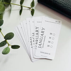 Peut inclure: Un ensemble de 12 cartes blanches avec du texte noir, chaque carte représentant un mois de l'année. Les cartes présentent un défi d'épargne mensuel avec un total d'épargne de 90. Le texte sur la carte est "Faites ce que vous aimez et l'argent suivra".