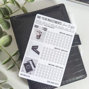 May include: A black faux-leather planner with a white savings tracker card. The card features the text "ONE-YEAR INVESTMENTS" and "Javan Budgets". The card has circles for tracking savings, with the text "EACH CIRCLE: £25" and "TOTAL SAVED: £3,375".