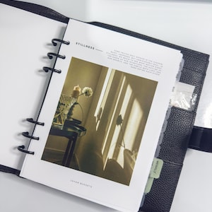 May include: Black leather planner with a printed page featuring the word "Stillness" and a photograph of a room with sunlight streaming through a window. The planner has black metal rings and labeled dividers.