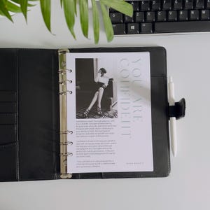 May include: A black leather binder with a white pen and a printed page that reads "YOU ARE CONFIDENT". The page features a black and white photo of a woman and text about confidence. The binder is open on a white surface.