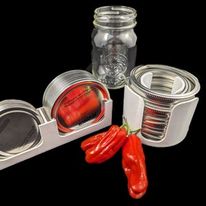 May include: A clear glass mason jar and several metal jar lids with white holders are displayed. Two red peppers are in the foreground. The lids have the text "crafted for quality sealed for freshness".