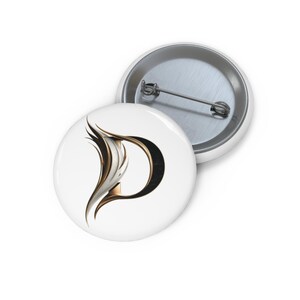 Op de afbeelding: Een ronde button met speld aan de achterzijde, voorzien van een gestileerde letter D op een witte achtergrond. De letter is uitgevoerd in een metallic goud en zwart kleurenpalet met zwierige, vederachtige lijnen aan de linkerzijde.