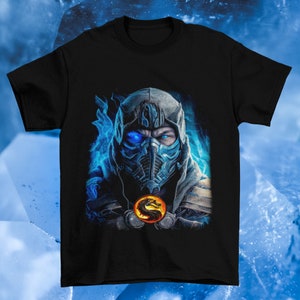 Puede incluir: Una camiseta negra con un gráfico de Sub-Zero de Mortal Kombat. El gráfico presenta la cara de Sub-Zero con sus característicos ojos azules y máscara, rodeada de llamas azules. El logotipo de Mortal Kombat es visible en el centro del gráfico.