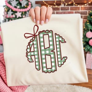 May include: Cream-colored t-shirt featuring a green and white plaid monogram design with the letters A, B, and I. The monogram is outlined in a dark red color and adorned with a matching bow at the top. The shirt is held up by a person.
