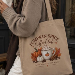 Könnte beinhalten: Eine beigefarbene Stofftasche mit einem grafischen Aufdruck einer Kaffeetasse mit Schlagsahne, Kürbissen, Zimtstangen und Kaffeebohnen. Der Text lautet PUMPKIN SPICE Coffee Club sowie COFFEE TODAY HAPPY ALWAYS, ergänzt durch kleine Herzsymbole.
