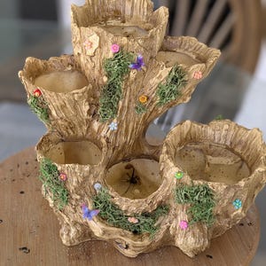 May include: A multi-tiered, tree trunk-shaped planter with several openings for plants. The planter is a light brown color with a textured surface, decorated with small, colorful flowers, green moss, and a butterfly. The planter is designed to hold multiple small plants.