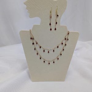 Può includere: Un set composto da una collana a doppio filo color oro e orecchini pendenti abbinati. La collana presenta due catenine delicate decorate con piccoli ciondoli a forma di goccia di colore rosso scuro. Gli orecchini sono composti da sottili catenine.