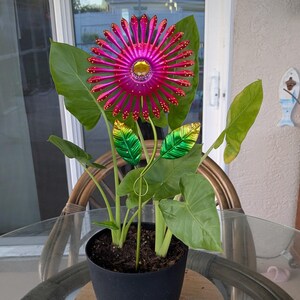 May include: A decorative metal flower with a magenta centre and petals, accented with gold and red details. The flower is planted in a black pot with large green leaves. The flower is a garden ornament.