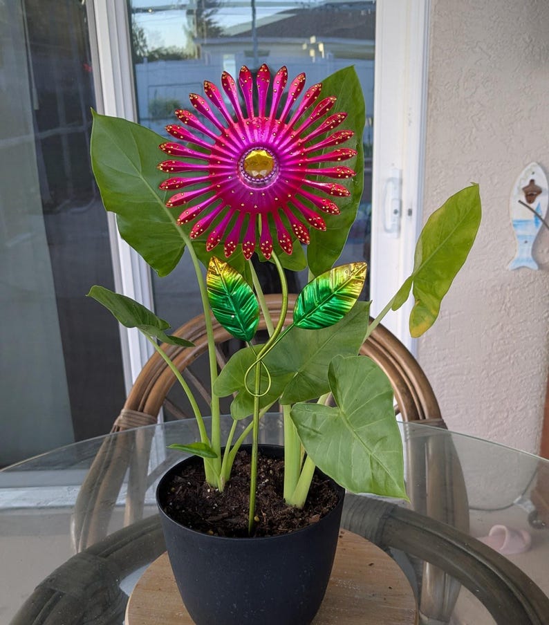 May include: A decorative metal flower with a magenta centre and petals, accented with gold and red details. The flower is planted in a black pot with large green leaves. The flower is a garden ornament.