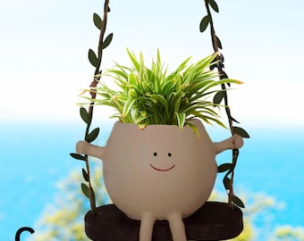 May include: A whimsical, off-white ceramic planter shaped like an egg with a smiling face, arms, and legs. The planter is suspended from a swing made of dark brown wood and vines. The planter holds a vibrant green plant with white stripes. The letter "C" is in the lower left corner.