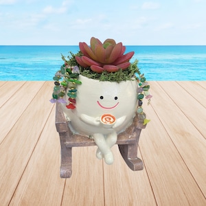 May include: A whimsical white ceramic planter shaped like a smiling character, holding a cup of coffee, and seated in a miniature rocking chair. The planter is filled with a succulent and decorative stones. The background shows a beach scene.