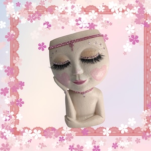 May include: A face-shaped planter with closed eyes, long eyelashes, and a pink jeweled headband and necklace. The planter has a neutral skin tone with pink blush and lips. The background is a pink floral design.