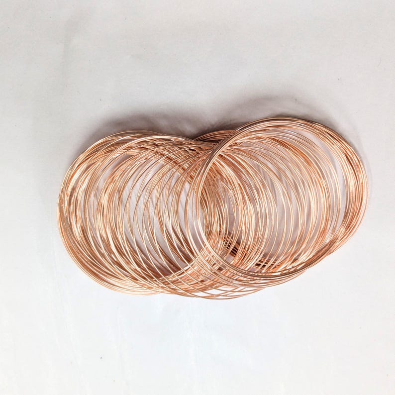 May include: A collection of thin, circular copper-colored wire bangles arranged in an overlapping, layered formation. The metallic rings feature a smooth, polished finish and a consistent, slender gauge, creating a dense, circular cluster.
