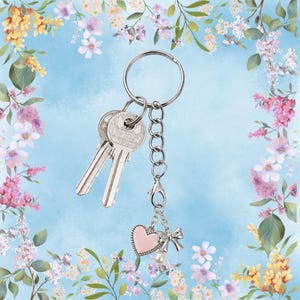 May include: Silver keychain with two keys and a pink heart charm with a bow and pearl accent. The keychain is attached to a chain and clasp. The background is a light blue with floral accents.