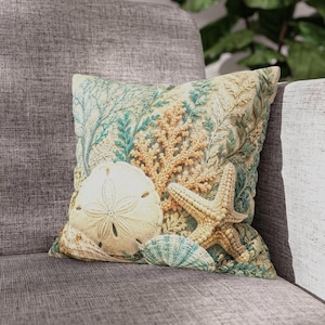 May include: A square throw cushion featuring a detailed print of marine life, including a light beige sand dollar, a textured tan starfish, and a blue-green seashell, set against a background of intricate coral and seaweed patterns in soft tones.